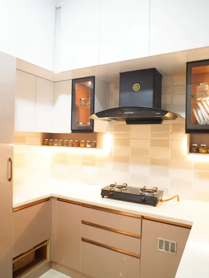 This L-shaped kitchen in a 3BHK apartment features beige cabinets with rose gold profile handles and a white countertop. The design includes glass-fronted display cabinets with internal lighting for an elegant touch.