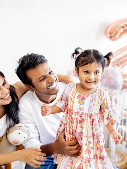 A happy family moment celebrating a second birthday at home. The balloons in the background add a festive touch, but the real story is the shared laughter between the parents and their daughter.