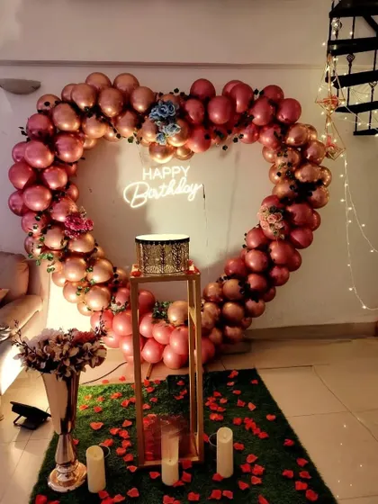 A romantic rose-gold balloon heart arch for an indoor birthday celebration. The setup is enhanced with flower bouquets and candles for an extra touch of elegance.