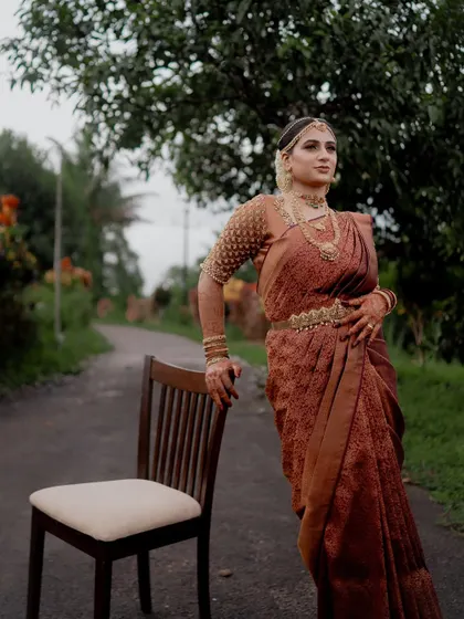 A portrait of the bride in her traditional saree, standing next to a chair on a rustic path.