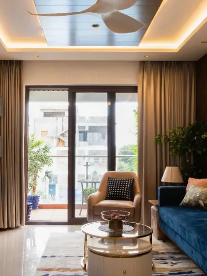 A full view of this living room, showcasing the deep blue sofa, modern TV unit with open shelving, and a false ceiling with reflective panels and cove lighting.