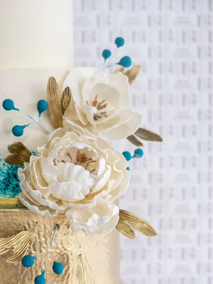 A detailed shot of our modern wedding cake, highlighting the delicate, handcrafted sugar peonies and the pop of color from the turquoise berries against the brushed gold and white tiers.