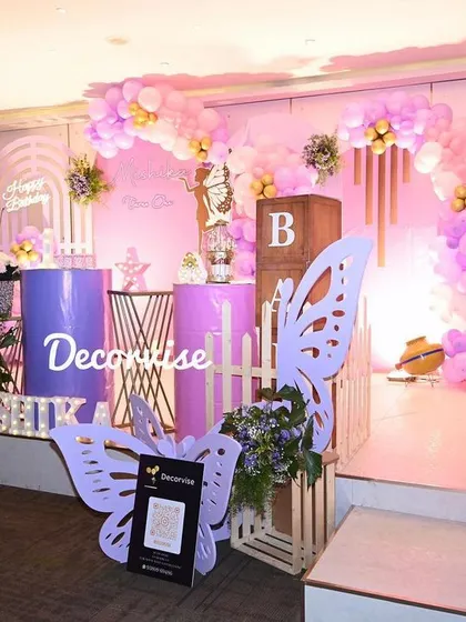 This wide view shows the complete butterfly-themed setup for Mishika's party. The stage includes a main backdrop with a balloon arch, a milestone board detailing her first year, and decorative props like wooden crates and a QR code stand for guests.