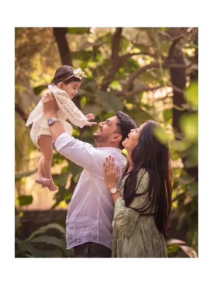 A joyful family photo in a park, with the father lifting his one-year-old daughter high in the air.