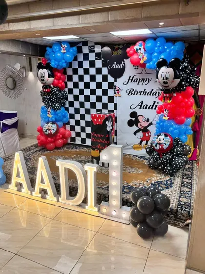 A classic Mickey Mouse first birthday party, featuring a checkered backdrop, balloon columns with Mickey foils, and large light-up letters for the name 'AADI'.