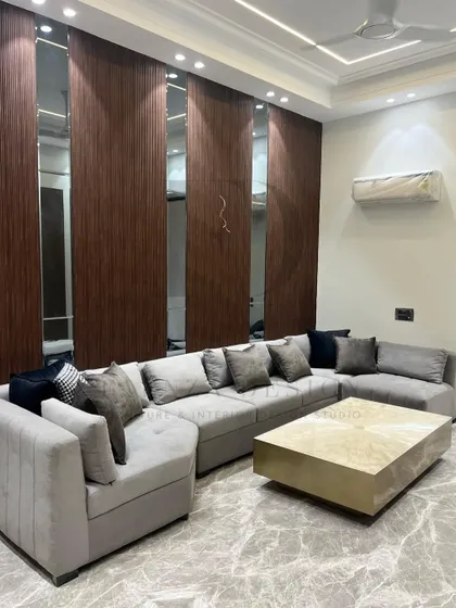 This L-shaped sectional sofa in a soft grey fabric provides ample seating, paired with a striking brass-finished center table. The back wall features a modern design of wood veneer and mirror panels, adding depth to the room.