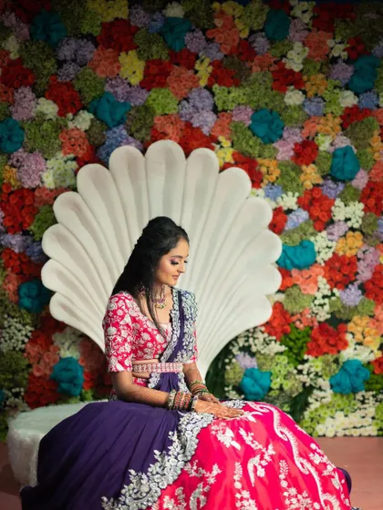 The bride seated against a stunning backdrop for her Mehendi. The seashell-shaped seat is set against a dense wall of multi-colored flowers, creating a vibrant and picture-perfect moment.