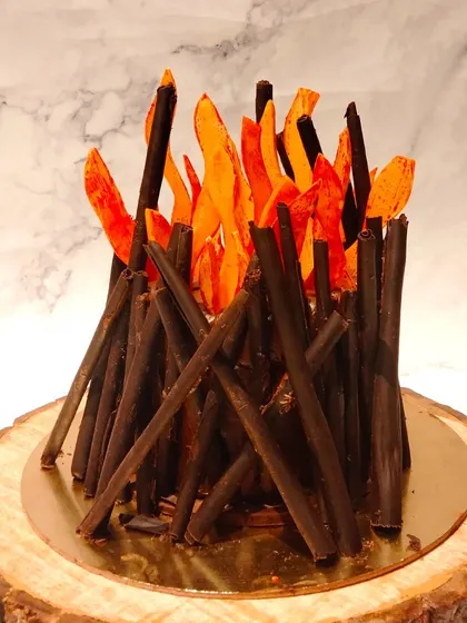 A close-up of the Lohri bonfire cake. The chocolate sticks are carefully arranged to resemble logs, and the caramelized sugar flames add a beautiful, warm glow.