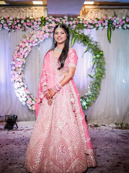 A full-length shot of the bride in her stunning pink lehenga, standing before the floral ceremony arch. This portrait captures her complete look and the beautiful decor of the event.
