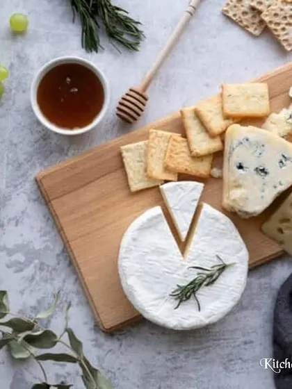 A deconstructed, top-down view of a classic cheese board. This flat-lay shot highlights a wheel of Brie, blue cheese, and hard cheese, accompanied by crackers, grapes, and a bowl of honey.