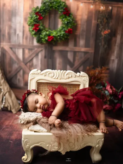 A sleeping baby girl in a rich red dress, resting on an elegant chaise against a rustic barn door backdrop. The Christmas wreath adds a festive touch.