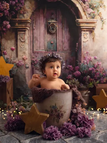 A magical, fantasy-themed sitter session. This baby girl is sitting in a pot surrounded by purple flowers, with a fairy-tale door in the background.