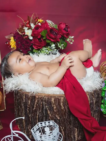 A happy baby lying on a log, draped in a red cloth, surrounded by flowers.
