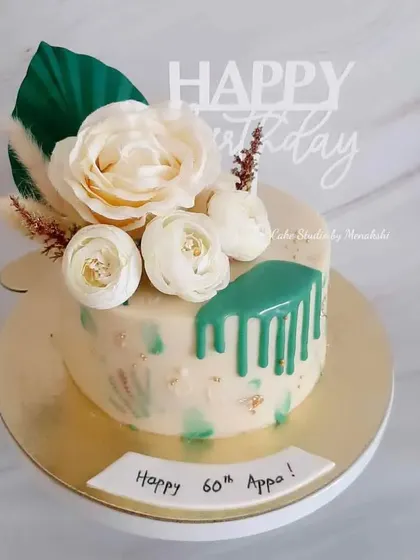 A milestone 60th birthday cake for Appa. This no-fondant design features a green drip effect and is adorned with white flowers and a dried palm leaf for a modern, natural look.