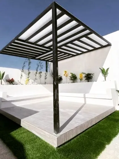 A minimalist white-and-black pergola on a raised platform. The design features a simple black metal frame with a translucent roof, set against a clean, multi-level white planter wall.
