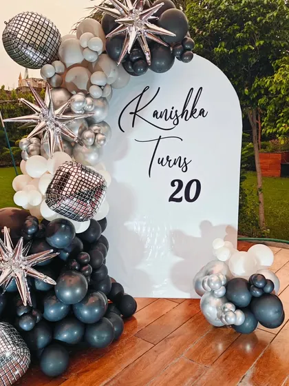 An outdoor view of the chic 20th birthday setup. The black, white, and silver balloon arch on the custom backdrop looks stunning against the greenery.