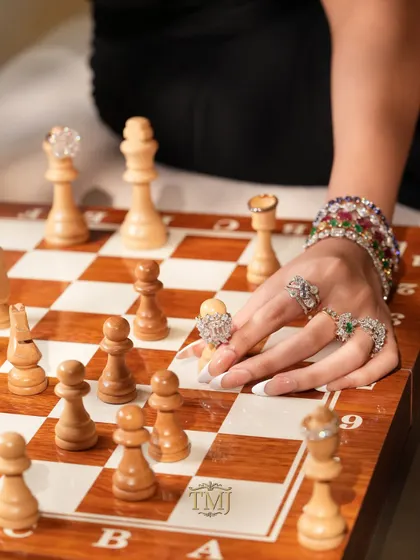 A close-up of a hand adorned with statement rings making a move on a chessboard, where each chess piece is also topped with a diamond ring.