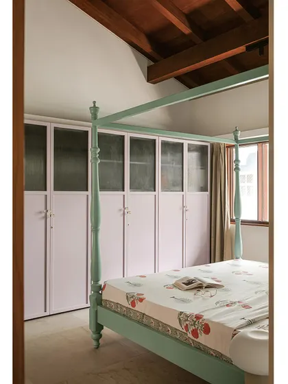 The bedroom features lilac-colored wardrobes with fluted glass, creating a soft contrast with the mint bed and the warm wooden beams overhead.