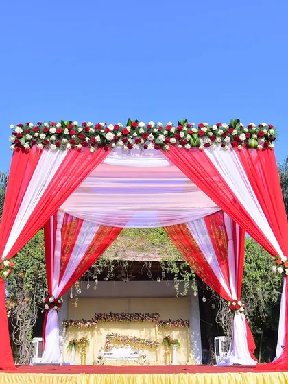 A stunning red and white mandap for an outdoor wedding. The bright colors stand out beautifully against the clear blue sky and lush greenery, creating a picture-perfect ceremony spot.