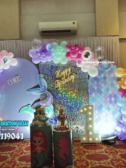 A mermaid-themed first birthday with a unique backdrop. The setup includes two panels, one with a shimmer wall and one with a mermaid scale print, connected by a pastel balloon arch.