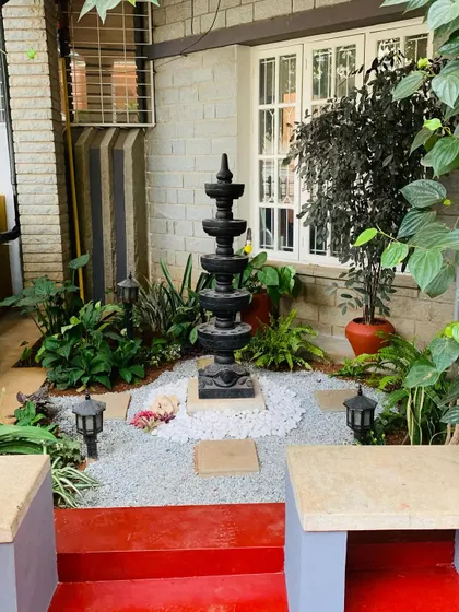 A wider view of the entrance garden featuring the Deepa Stambha. The mix of dark and light foliage, along with the traditional elements, creates a welcoming and culturally rich entryway.