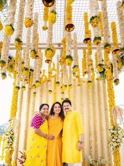 A happy family posing in front of a haldi backdrop I designed. The ceiling is covered with hanging strings of white and yellow flowers with small bells, creating a beautiful and festive atmosphere.