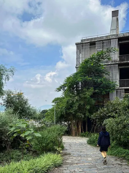 A student walks along a stone path at Avasara, flanked by mature trees and dense shrubbery. The pathways are designed to meander through the landscape, encouraging exploration and connection with nature.