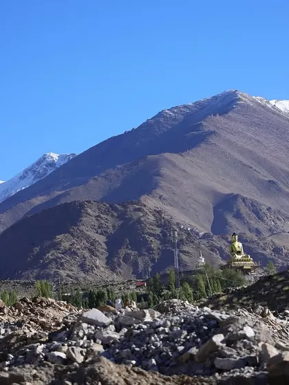 A golden Buddha statue sits peacefully in the valley, with the rugged, snow-tipped mountains rising behind it.