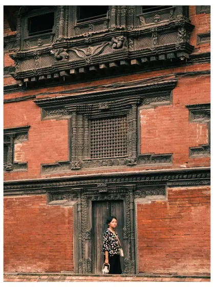 A woman stands in the doorway of an ancient building in Kathmandu, surrounded by intricate brickwork and carved wooden windows. The shot blends portraiture with architectural documentation.