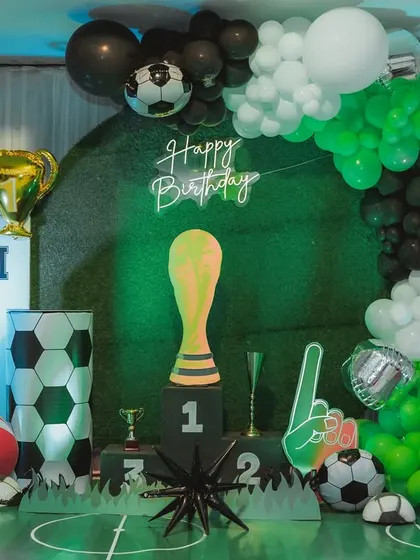A full view of a football-themed birthday stage. The setup includes a turf-like floor, a winner's podium, a giant trophy prop, and cutouts of famous football players.