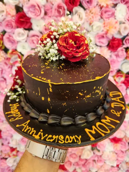 A rich, dark chocolate anniversary cake for 'Mom & Dad', decorated with red roses and a gold splatter effect.