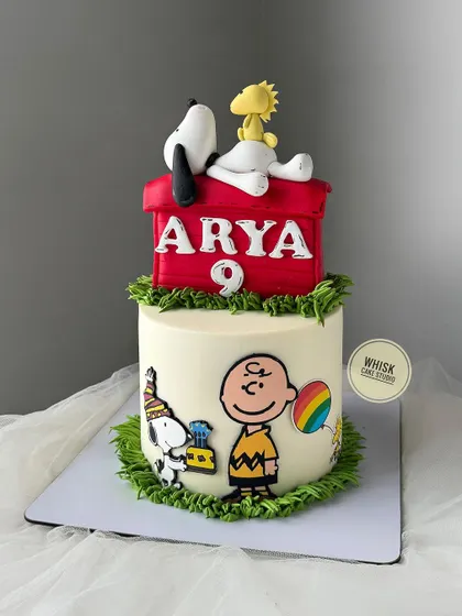 A charming Snoopy-themed cake. This design features Snoopy and Woodstock relaxing on top of their iconic red doghouse, with hand-painted details on the side.