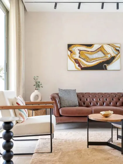 A living room from the House of Hiranandani project, featuring a classic brown leather sofa paired with a modern armchair. The abstract gold and black artwork adds a touch of drama to the neutral space.