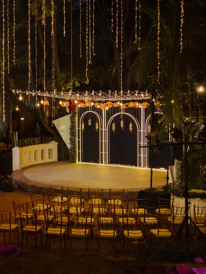 A stunning Sangeet stage setup in an outdoor amphitheater. The space is illuminated by strings of fairy lights and features a modern arched backdrop, setting the scene for a night of performances and celebration.