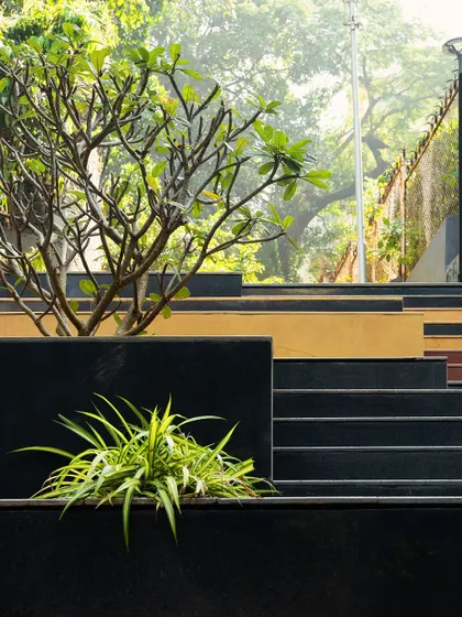 **Nepean Greens, Mumbai:** The design uses multi-level platforms and integrated planters to break up the space and create intimate nooks. The interplay of black granite, yellow Jaisalmer stone, and greenery adds visual interest and defines different zones.
