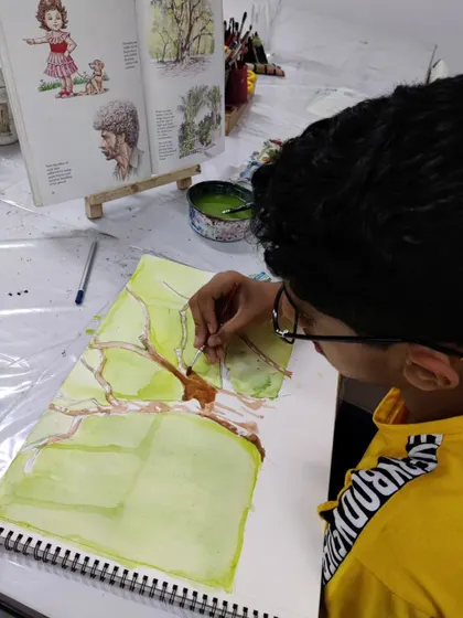 The student adds the trunk and branches to the watercolor wash, carefully defining the structure of the tree.