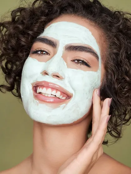 A playful and fresh image of a model applying a green clay mask. Her smiling face and direct eye contact create a fun and engaging visual. This shot is perfect for showing the application process of a mask in a way that feels enjoyable and part of a healthy lifestyle.