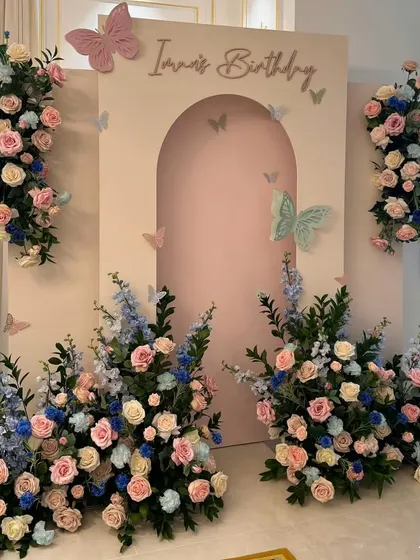 An elegant floral birthday setup with a pastel pink arched backdrop, surrounded by beautiful arrangements of pink, blue, and white flowers for a soft, dreamy look.