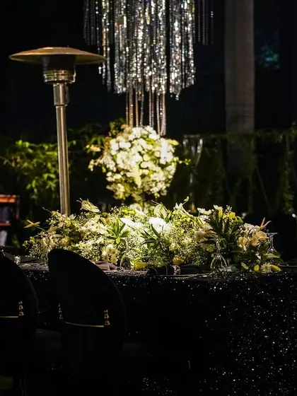 A table at a cocktail party, featuring a black sequined tablecloth, a centerpiece of white and green florals, and a modern tassel chandelier.