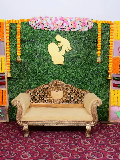 This backdrop combines a green artificial grass wall with a grid of marigold flowers and traditional decorative elements. A gold-colored ornate sofa completes the stage seating for the main guests.
