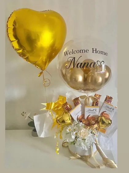 A golden "Welcome Home" hamper for Nana. This arrangement combines a personalized bubble balloon, a gold foil heart, and a bouquet filled with Lindt chocolates and white flowers.