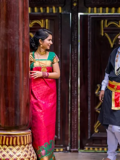 A playful, candid moment between pillars. The bride's bright saree and the groom's traditional Kodava attire create a beautiful contrast in this heritage setting.