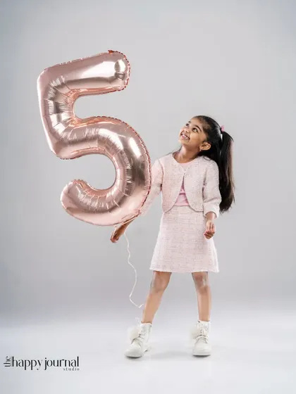 Celebrating five years! A girl in a chic pink outfit holds a large number '5' balloon, looking up at it with excitement.