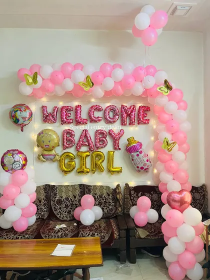 A 'Welcome Baby Girl' decoration for a home setting. The setup includes a pink and white balloon arch, foil balloons, and fairy lights over a sofa.