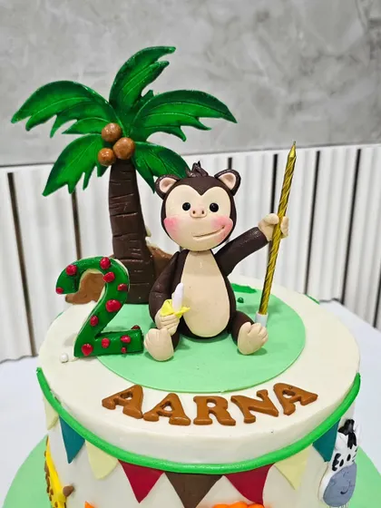 A cheerful jungle-themed cake for a second birthday. The topper features a playful monkey holding a banana, a custom nameplate, and a number '2' candle, all crafted from fondant.