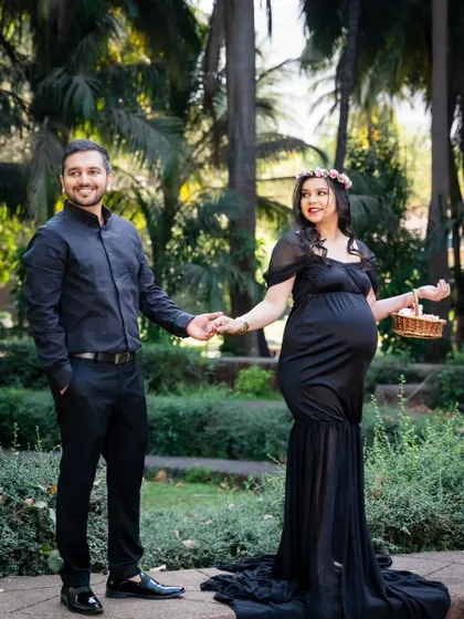 A full-length portrait of the couple in a beautiful garden. The mother-to-be's flowing black gown and flower crown add a touch of elegance to this outdoor maternity shoot.