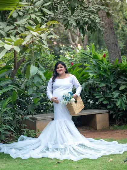 A serene portrait of a mom-to-be in a white gown, standing in a park and holding a basket with blue flowers.