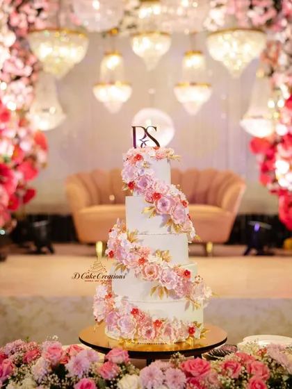 A classic love story calls for a timelessly romantic cake. This five-tier wedding cake features a cascading spiral of handmade sugar flowers in soft pinks and whites, accented with delicate gold leaves for a touch of luxury.