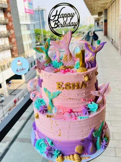 A vibrant two-tier mermaid theme cake for a first birthday. The cake is decorated with pink and purple buttercream, fondant mermaid tails, and golden seashells.