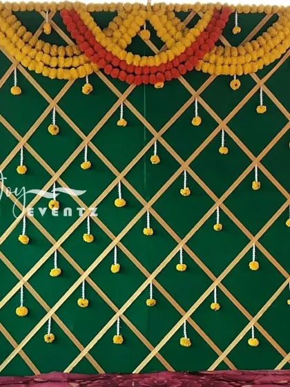 A traditional backdrop design with a green fabric base and a gold ribbon lattice pattern. The decor is framed with yellow marigolds and accented with small hanging floral bunches.